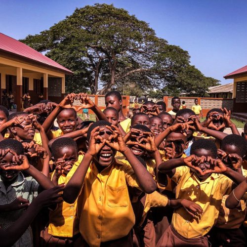 A large group of school children in yellow uniforms stand outside their school building, making heart shapes with their hands and smiling enthusiastically at the camera.