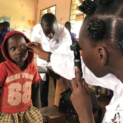 A healthcare worker examines a young child’s eye using a direct ophthalmoscope. The child, wearing a red hoodie, sits quietly while the examination is conducted.