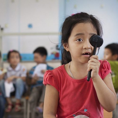 A young girl in a red shirt holds an occluder to her eye during an eye screening in a classroom, with other children sitting and waiting their turn in the background.