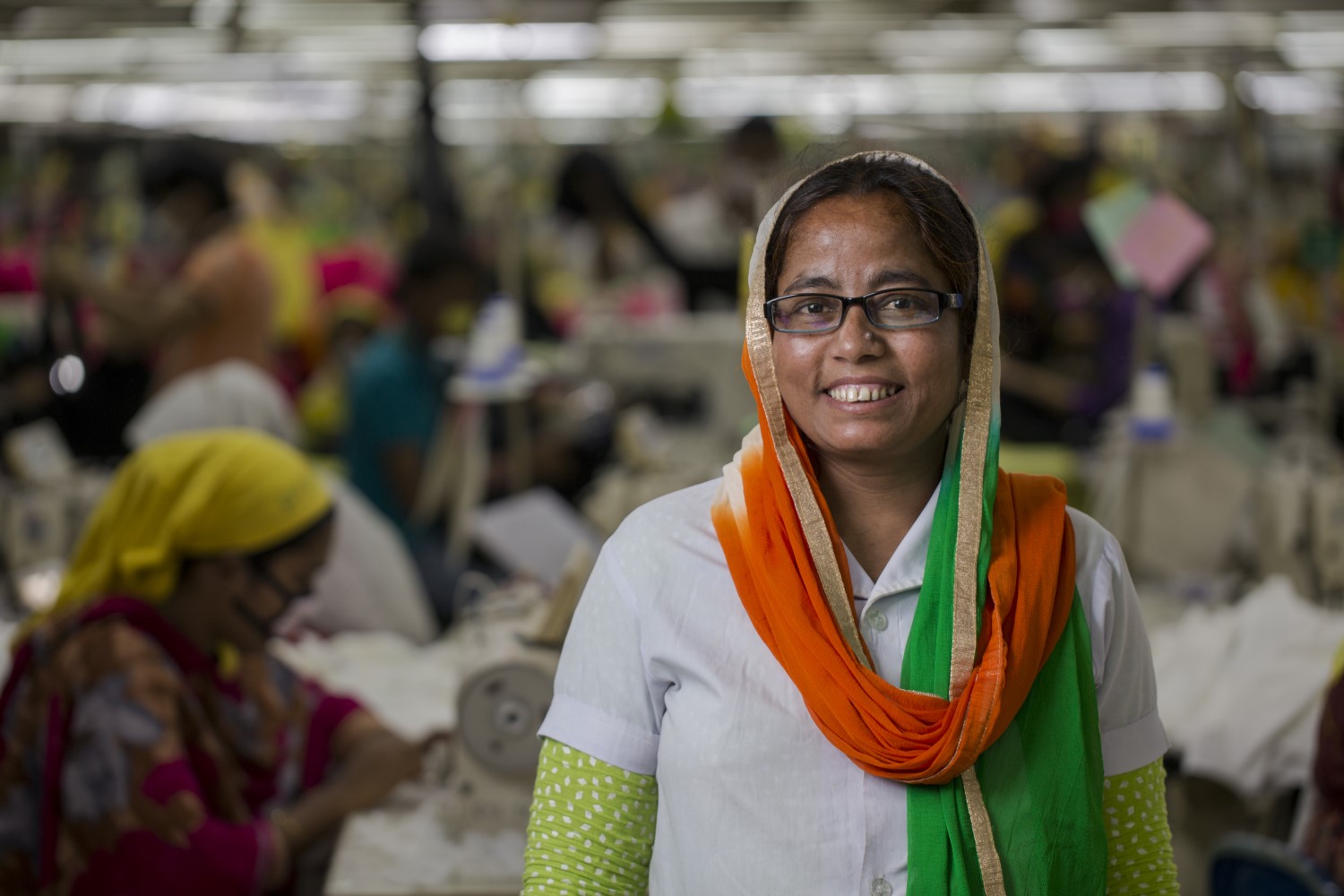 A woman wearing a headscarf and glasses smiles in a busy garment factory with other workers in the background.