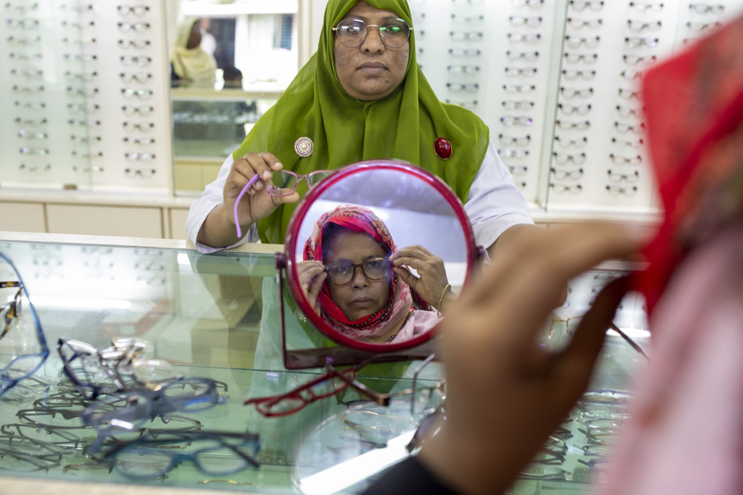 A woman in a green headscarf helps another woman, who is looking in a mirror, try on eyeglasses in an optometry store with multiple glasses on the counter and display shelves in the background.